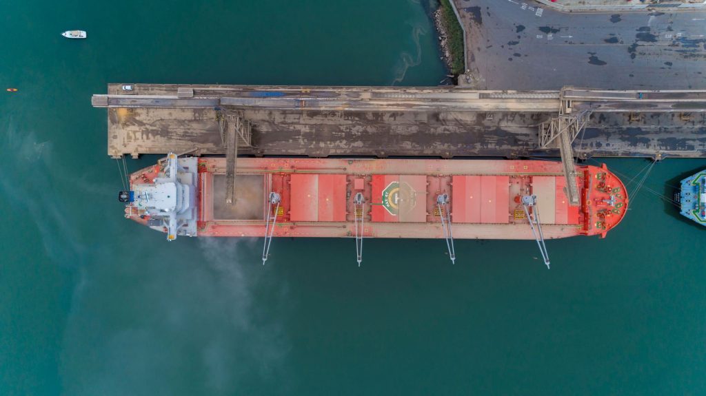 Portos brasileiros passam a ter política nacional de pesquisa e inovação Vista aérea de um navio cargueiro atracado em um porto brasileiro, com áreas de contêineres e estruturas de movimentação de cargas.