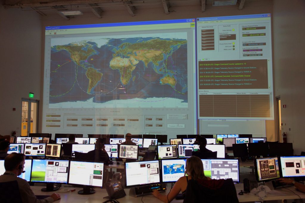 Equipe vista de costas analisando dados em computadores diante de um grande mapa digital em uma sala de controle.